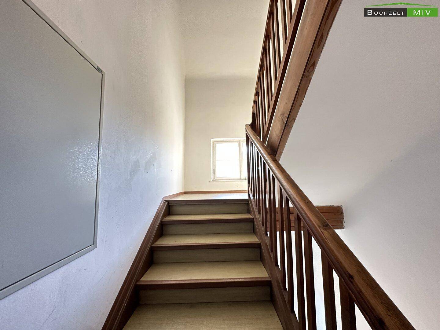 Holztreppe mit Geländer in einem hellen Treppenhaus, das zu den oberen Stockwerken führt.
