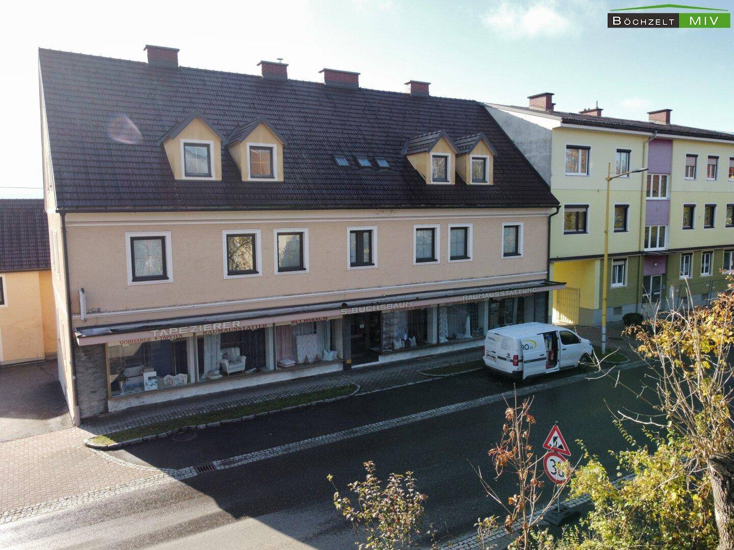 Mehrstöckiges Gebäude mit Geschäftsräumen im Erdgeschoss und Wohnungen darüber, an einer Straße gelegen.