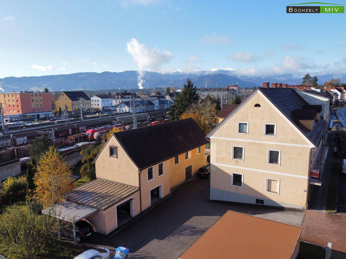 Luftaufnahme von mehreren Gebäuden mit Bahngleisen im Hintergrund und schneebedeckten Bergen am Horizont.