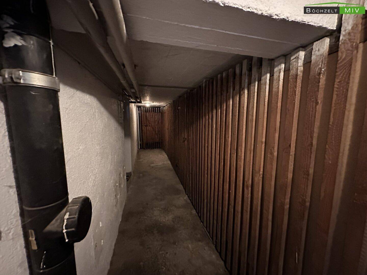 Langer Kellergang mit Holztrennwänden und Betonboden, der zu weiteren Lagerräumen führt.
