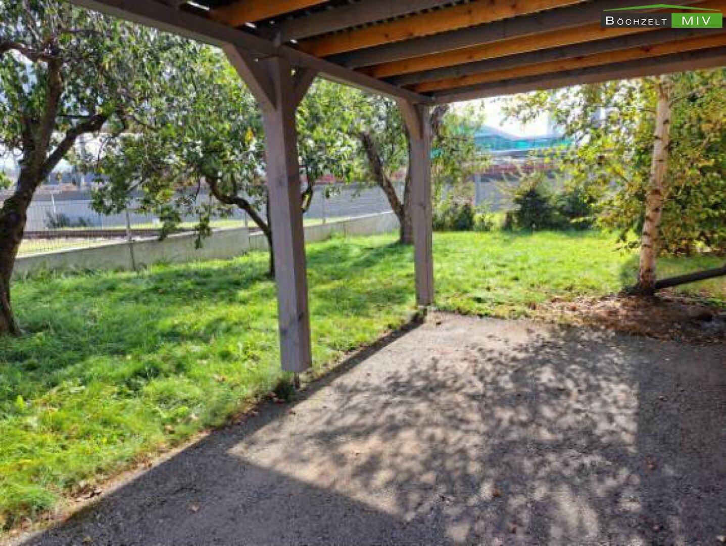 Überdachter Bereich im Garten, möglicherweise ein Carport oder eine Terrasse, umgeben von Bäumen.