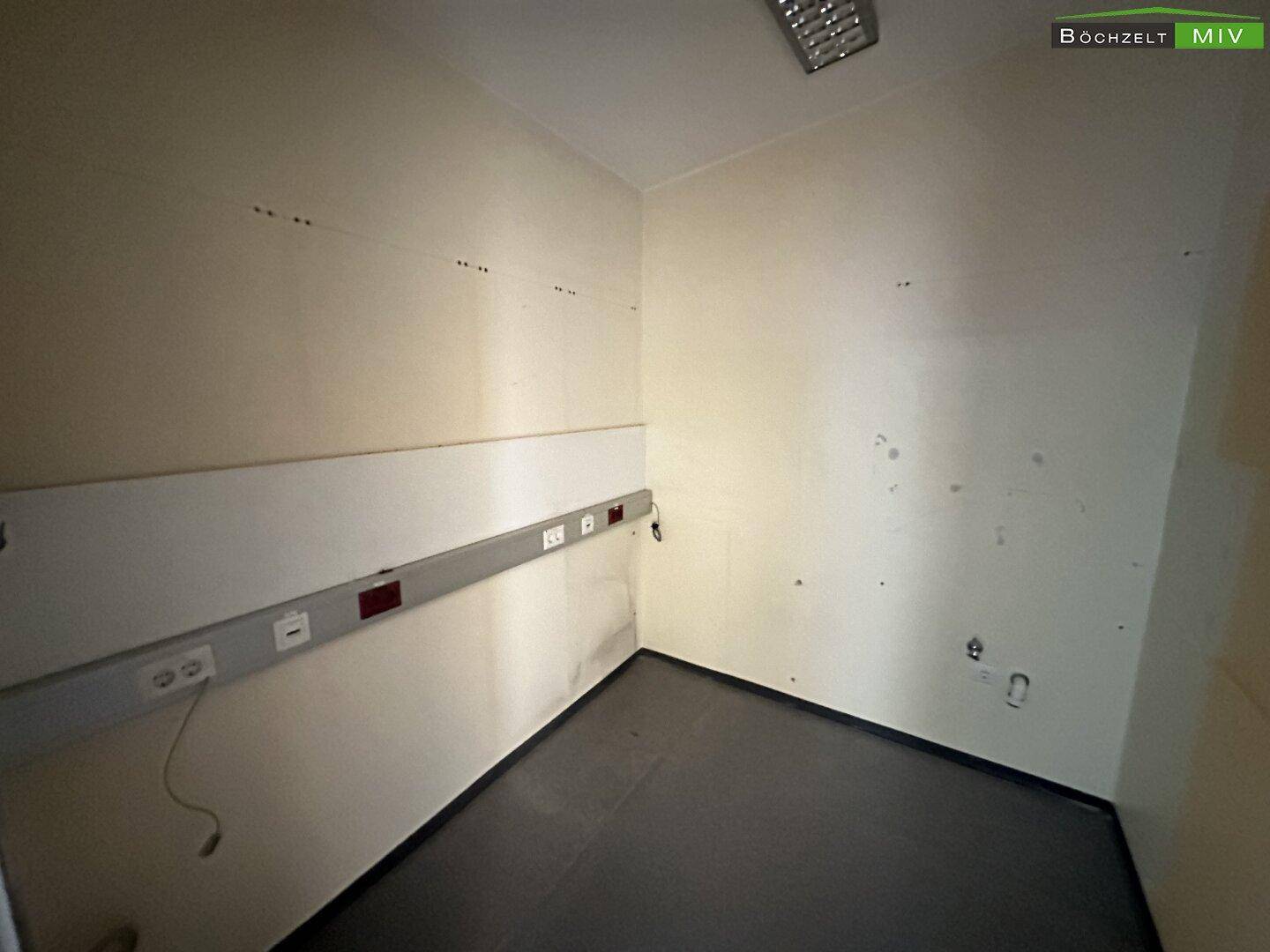 Leerer Raum mit dunklem Vinylboden und hellen Wänden, ausgestattet mit Steckdosenleisten und in gutem Zustand.