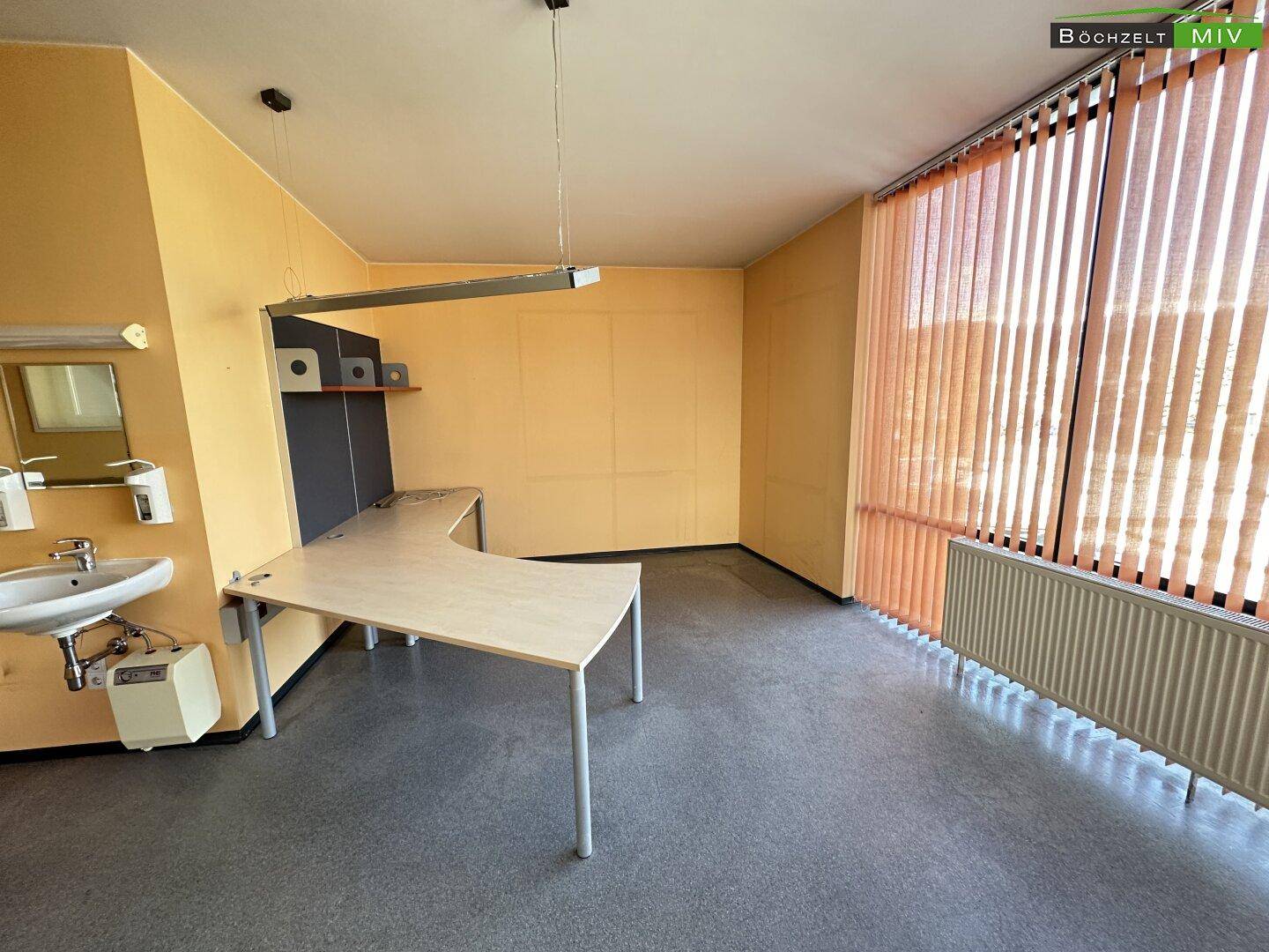 Geräumiges Büro mit Schreibtisch, Waschbecken und großen Fenstern mit Jalousien, die viel Tageslicht hereinlassen.