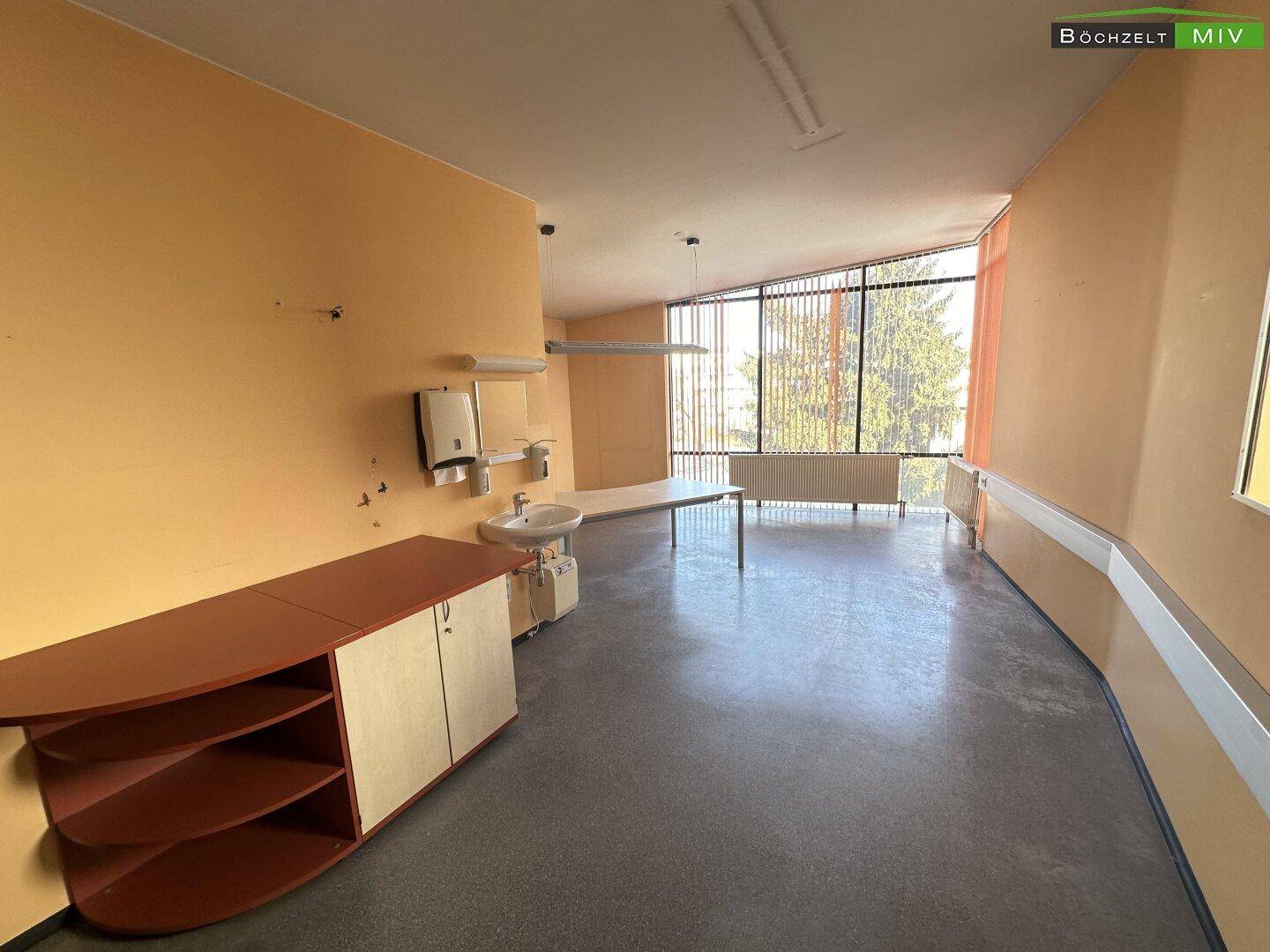 Großes Büro mit viel Tageslicht, Schreibtisch und Blick ins Grüne durch die Fenster mit Jalousien.