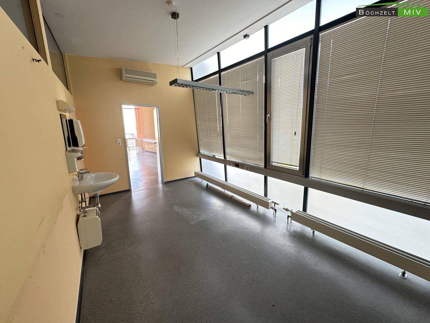Büro mit Klimaanlage, Waschbecken und großen Fenstern mit Jalousien, die für angenehmes Raumklima sorgen.