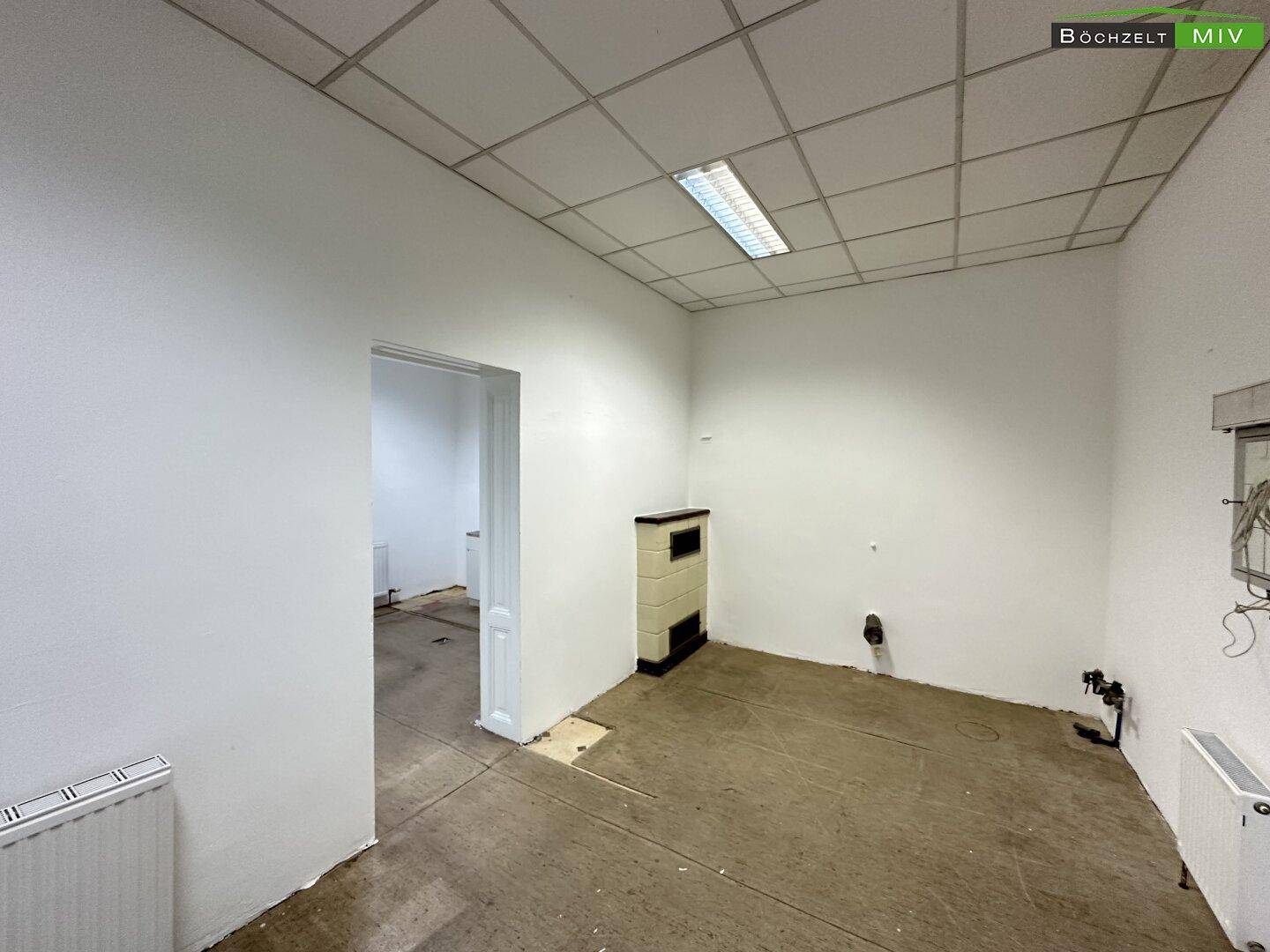 Leerer Raum mit Kachelofen, abgehängter Decke und sichtbaren Heizungsrohren, bereit zur Renovierung.
