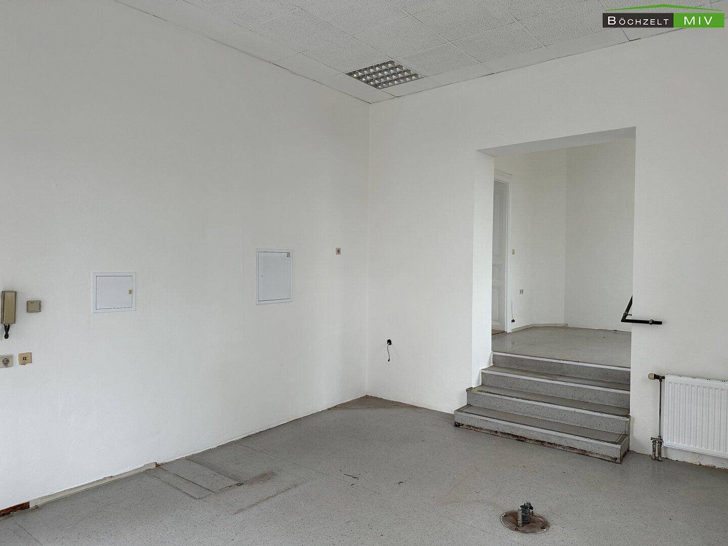 Leerer Gewerberaum mit hellen Wänden, abgehängter Decke und Stufen zu einem erhöhten Bereich.