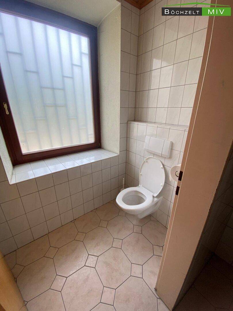 Einfache Toilette mit weißem Fliesenboden, Wand-WC und einem Fenster für Tageslicht.
