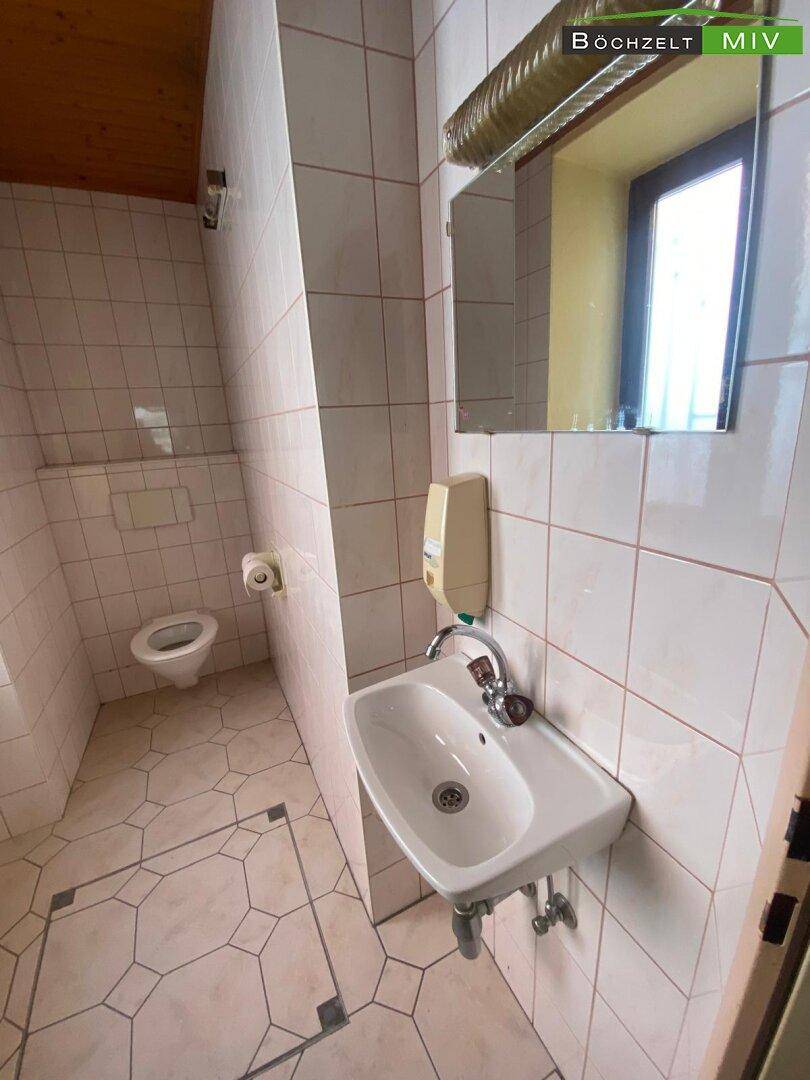 Toilette mit Waschbecken, Spiegel und Fenster, ausgestattet mit weißen Fliesen und Holzdecke.