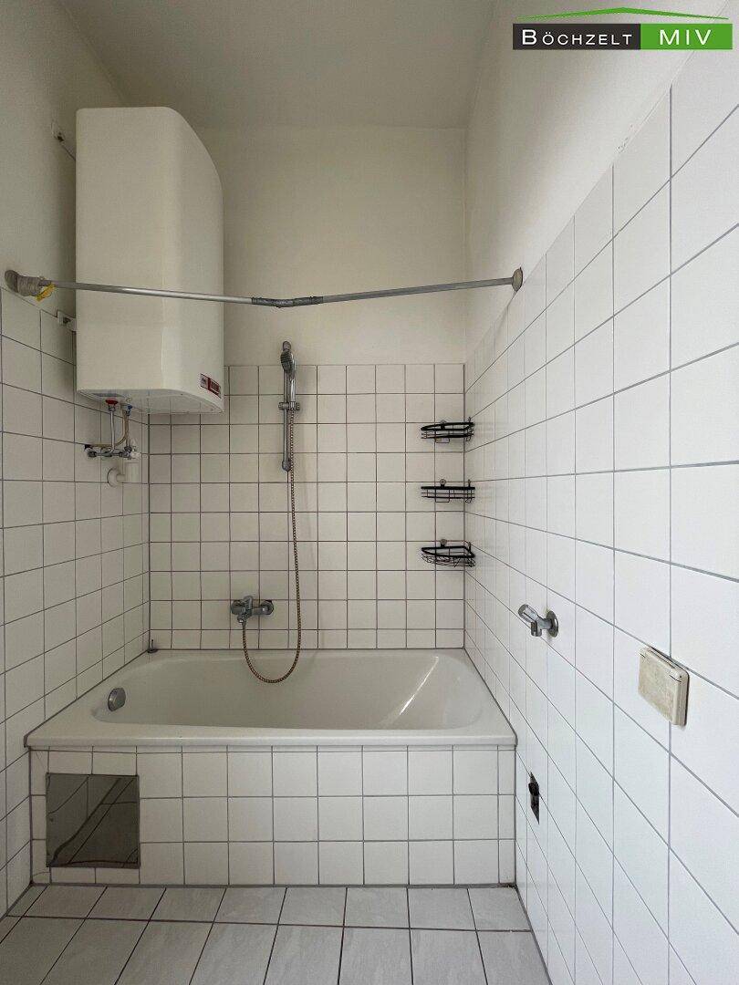 Gefliestes Badezimmer mit Badewanne und Duschvorrichtung, sowie einem Wandboiler für Warmwasser.