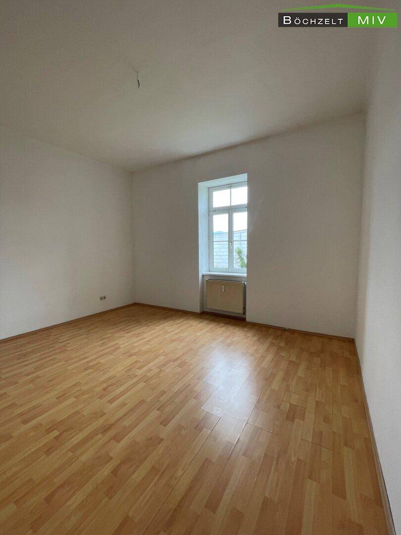 Helles, leeres Zimmer mit Laminatboden und einem Fenster, das viel Tageslicht hereinlässt.