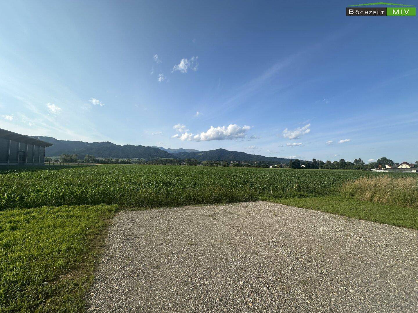 Blick über ein Kiesfeld auf weite grüne Felder und eine Bergkette am Horizont unter blauem Himmel.