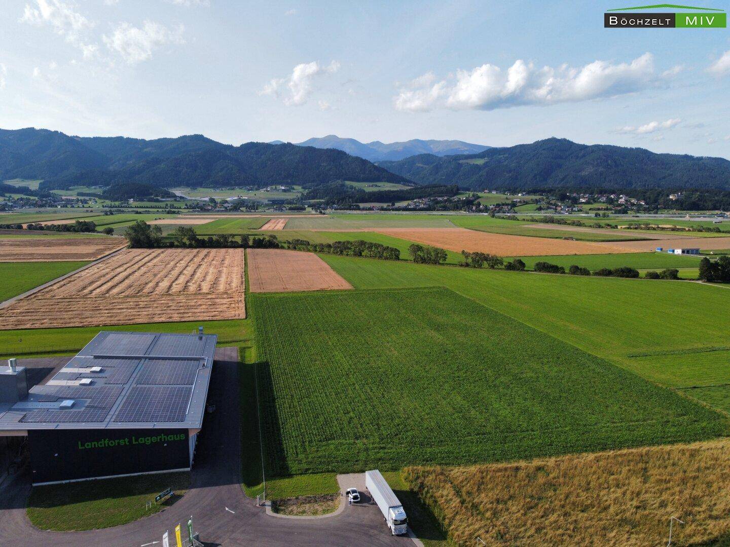 Luftaufnahme von Feldern und Bergen, mit einem Gewerbegebäude, das Solarpaneele auf dem Dach hat.