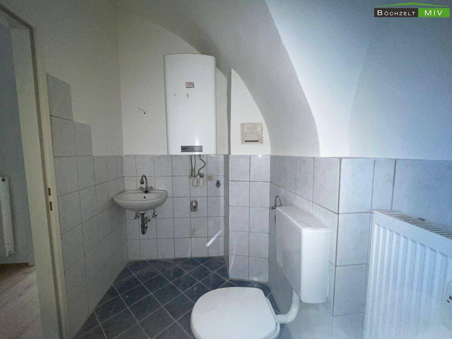 Badezimmer mit Toilette, Waschbecken und Boiler, ausgestattet mit weißen Wandfliesen und Dachschräge.
