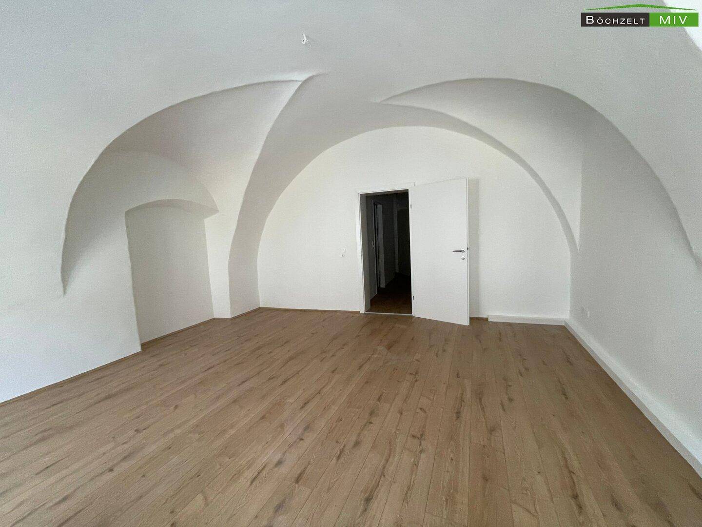 Großer Raum mit Holzboden und beeindruckender gewölbter Decke, eine offene Tür führt in einen Nebenraum.
