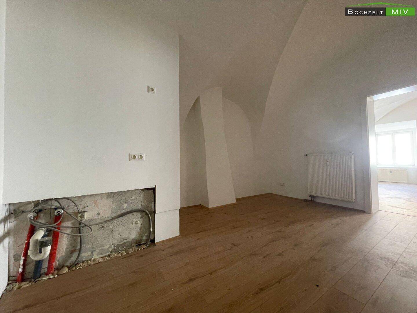 Raum mit Holzboden und vorbereiteten Anschlüssen für eine Küche oder ein Badezimmer, noch unfertig.