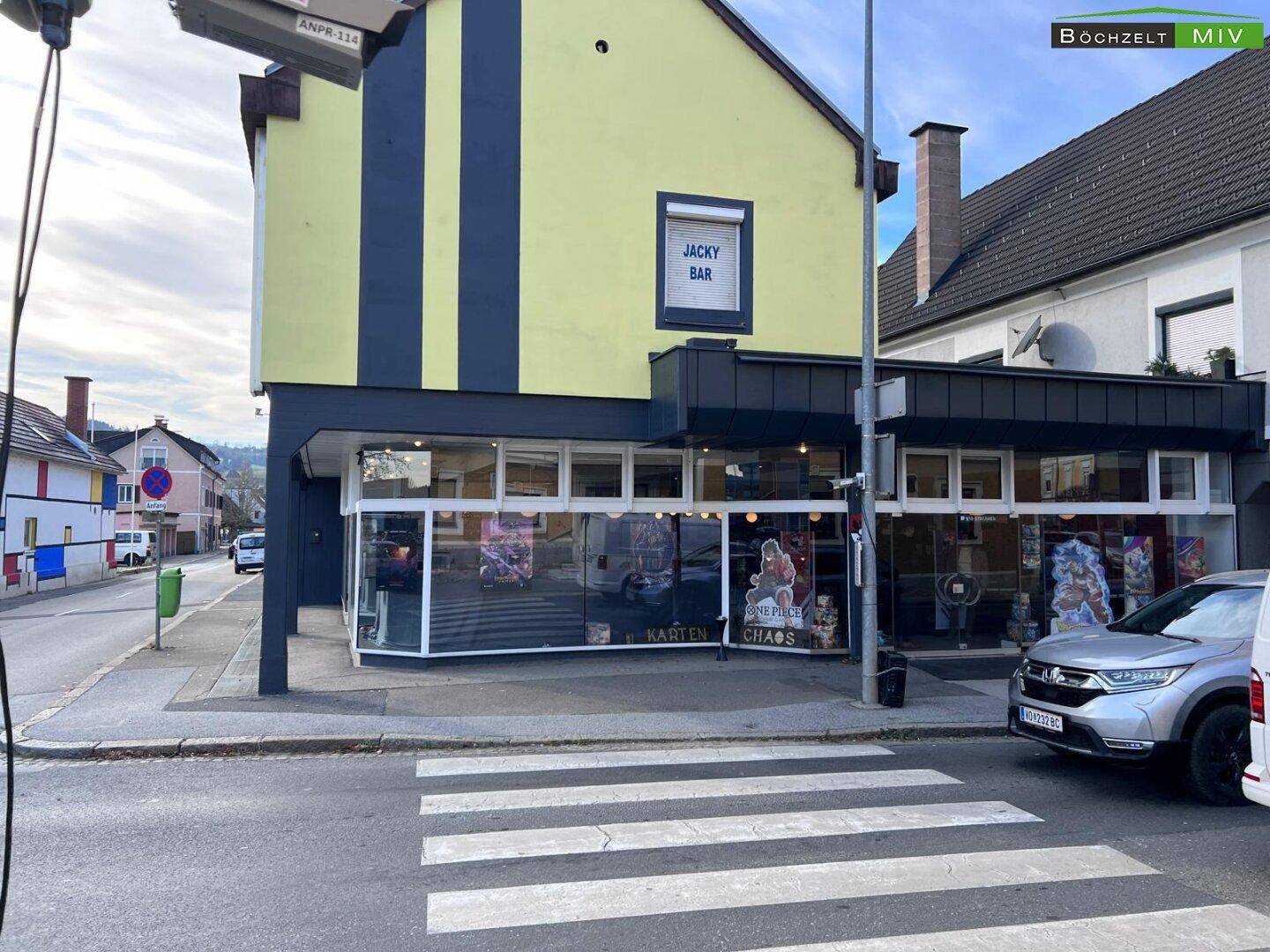 Seitenansicht des Gebäudes mit gelber Fassade, dunklen Streifen und großen Schaufenstern im Erdgeschoss.