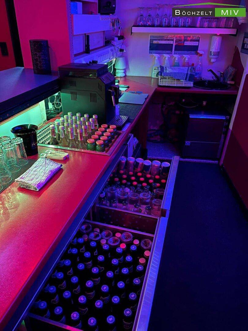Gut ausgestatteter Barbereich mit Flaschen, Gläsern und Kaffeemaschine unter roter Beleuchtung.
