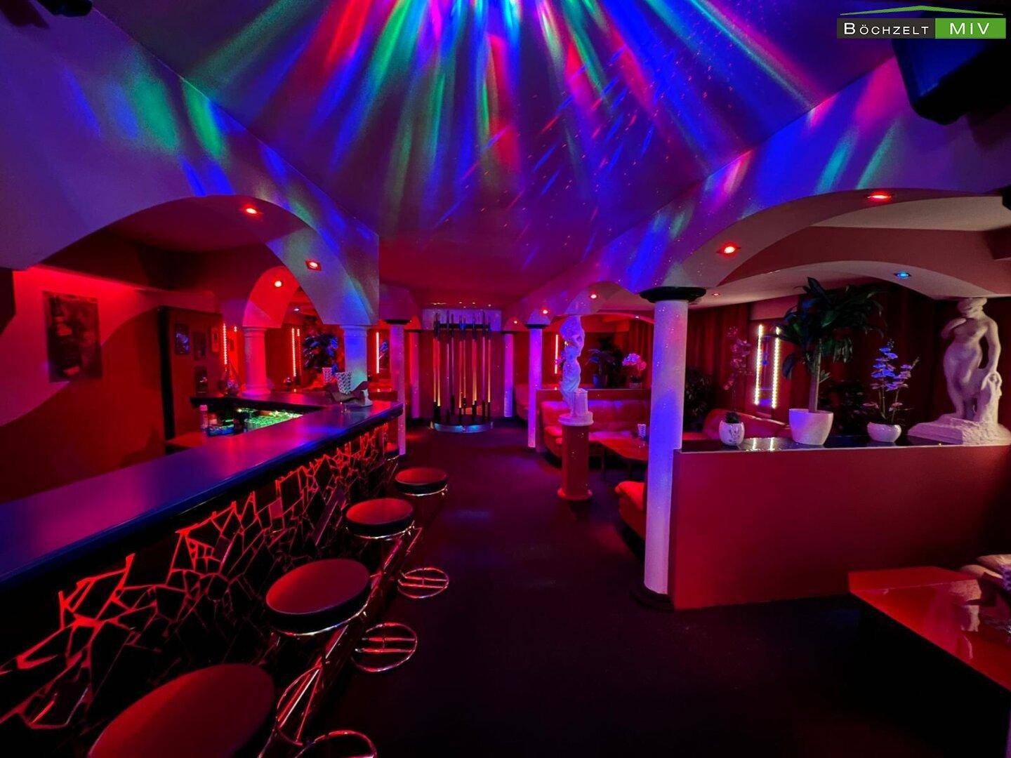 Hauptbarbereich eines Nachtclubs mit farbiger Deckenbeleuchtung, Barhockern und roten Akzenten.