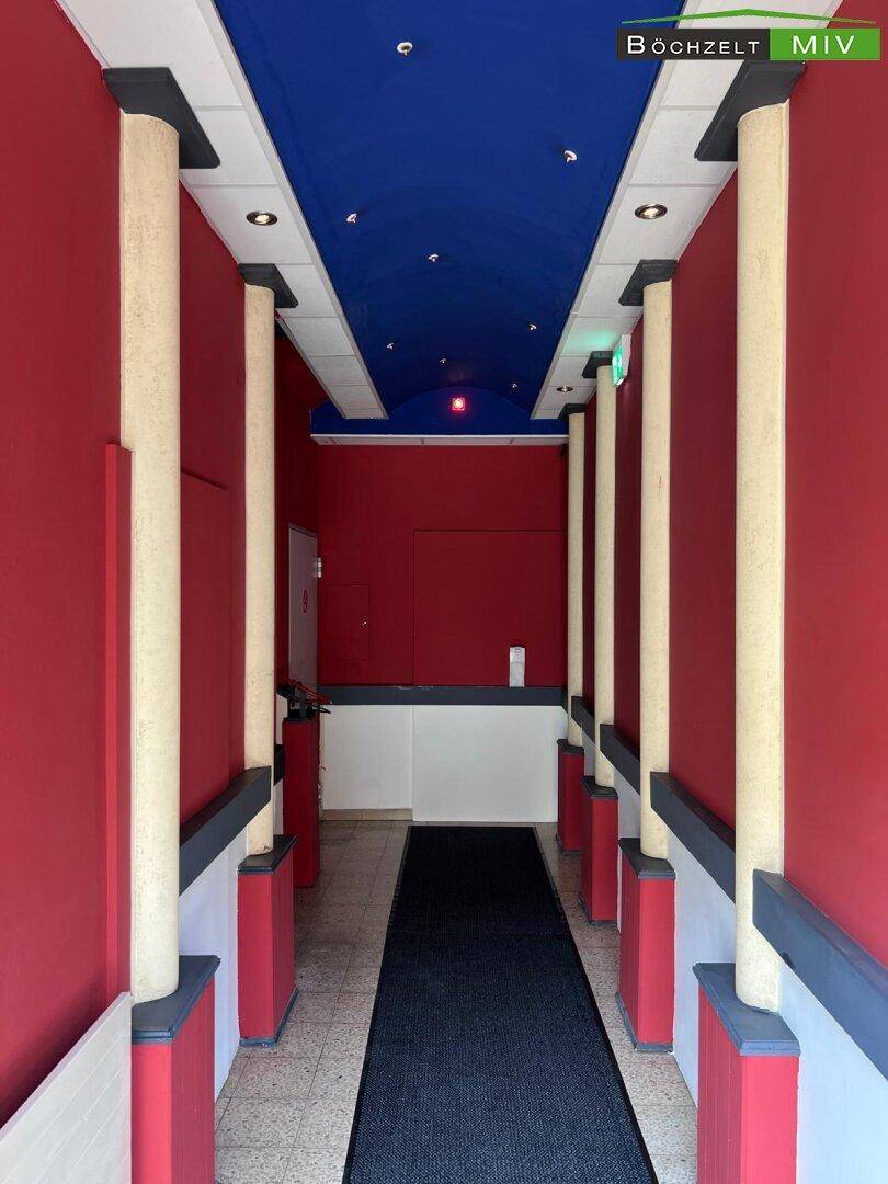 Langer Flur mit roten Wänden, blauer Decke und Säulen, der zu einem Eingangsbereich führt.