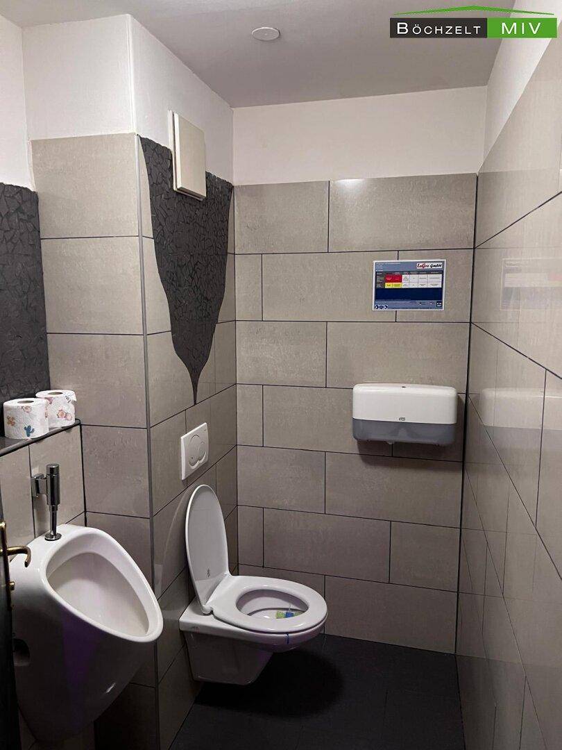 Moderne Gästetoilette mit Urinal, WC und grauen Fliesen an Wänden und Boden.