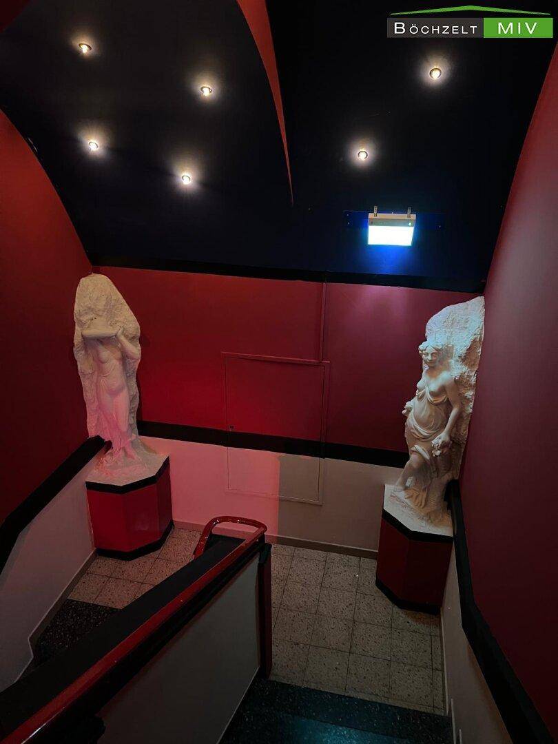 Treppenaufgang mit dunklen Stufen, roten Wänden und zwei klassischen Statuen als Dekoration.
