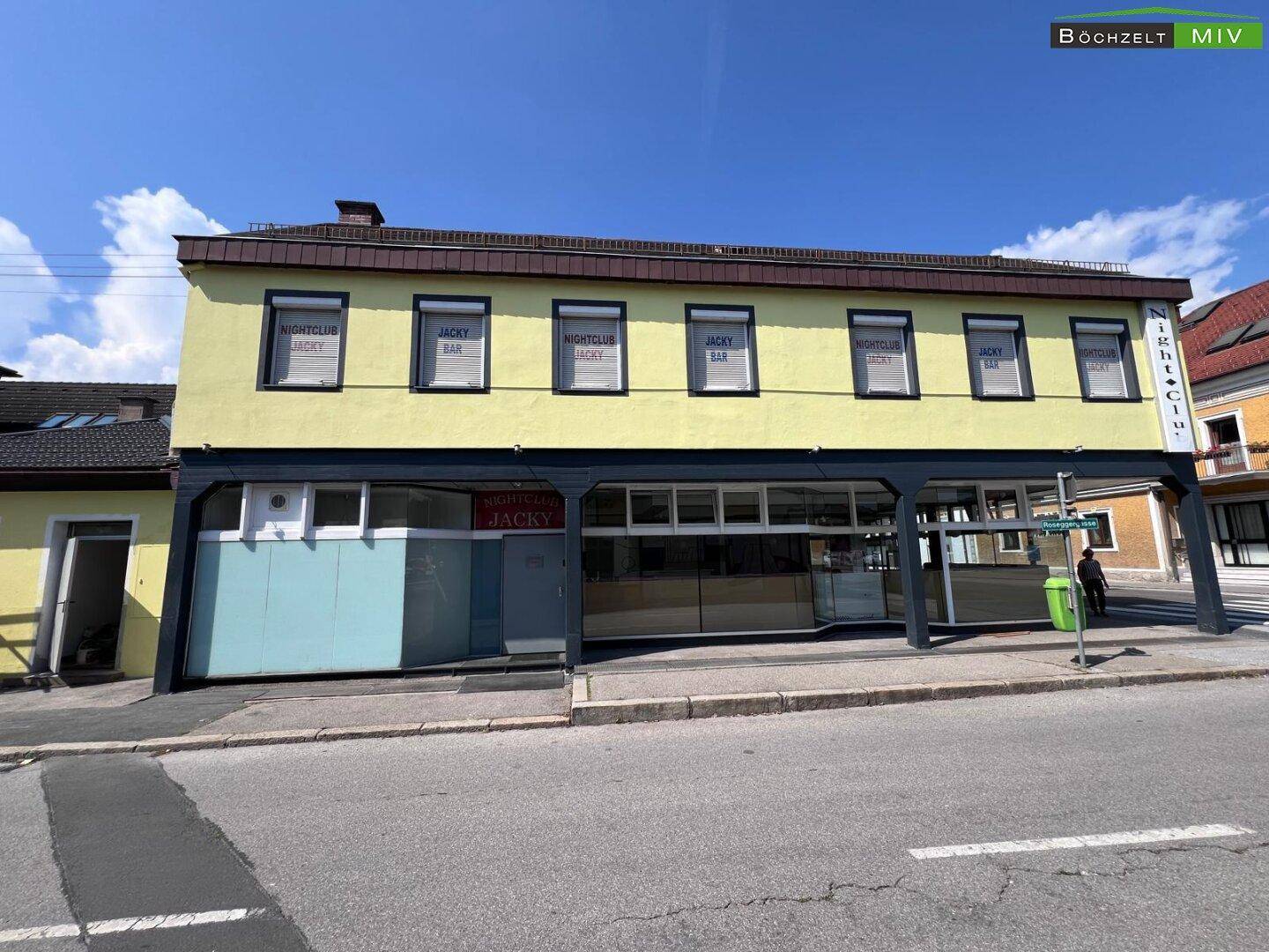 Gesamtansicht des Gebäudes mit gelber Fassade, dunklen Akzenten und großen Schaufenstern an einem sonnigen Tag.