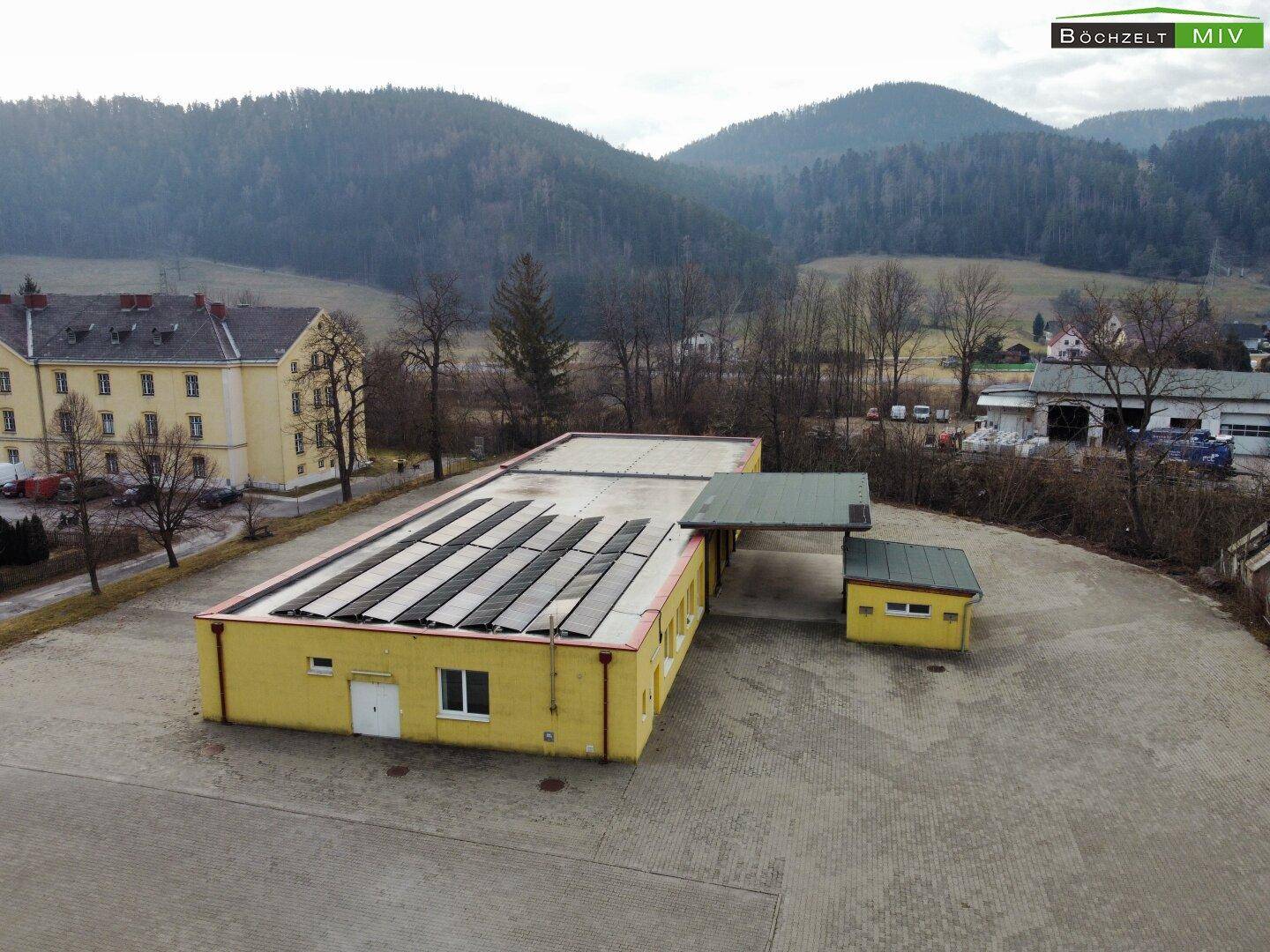 Gelbes Gebäude mit Solaranlage auf dem Dach, umgeben von einem großen Parkplatz und bewaldeten Hügeln.