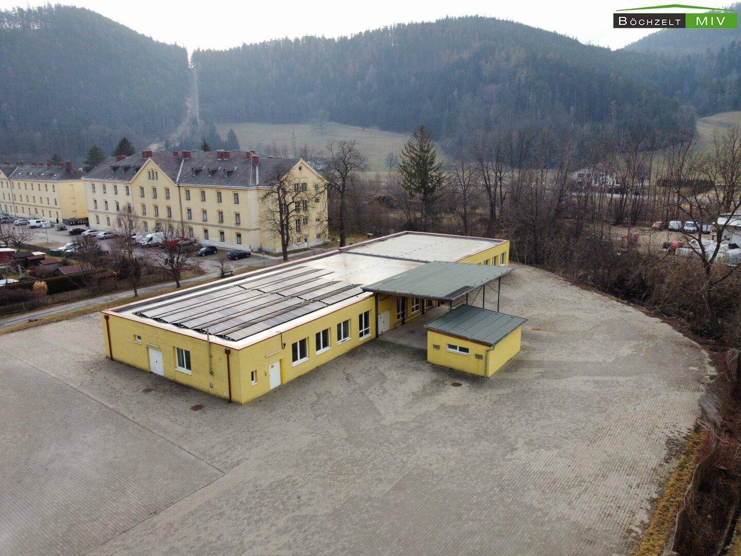 Luftaufnahme eines gelben Gebäudes mit Solarpaneelen auf dem Dach und einem größeren Gebäude im Hintergrund.