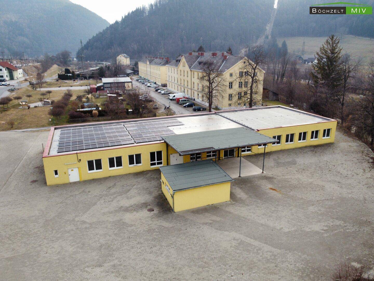 Vogelperspektive eines gelben Gebäudes mit Solarpaneelen und einem überdachten Eingangsbereich, umgeben von einer weiten Fläche.