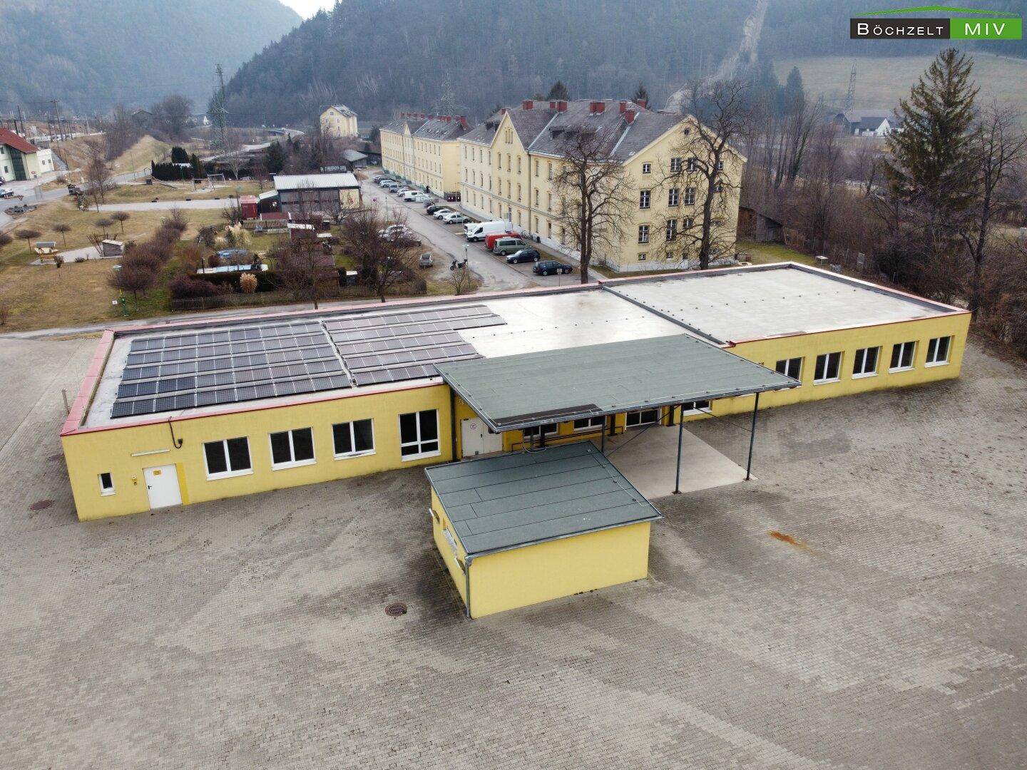 Luftaufnahme eines langgestreckten gelben Gebäudes mit Solaranlage und einem kleineren Anbau, im Hintergrund Berge.