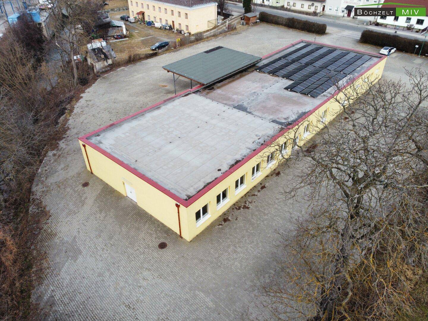 Luftaufnahme eines gelben Gebäudes mit Flachdach und Solarpaneelen, umgeben von einem großen gepflasterten Bereich.