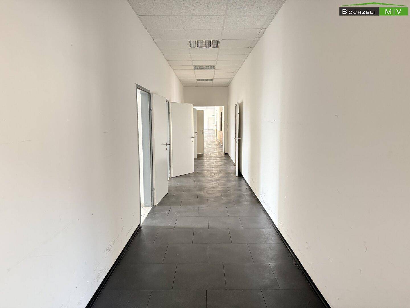 Langer, leerer Flur mit dunklen Fliesen und weißen Wänden, mehrere Türen führen in angrenzende Räume.