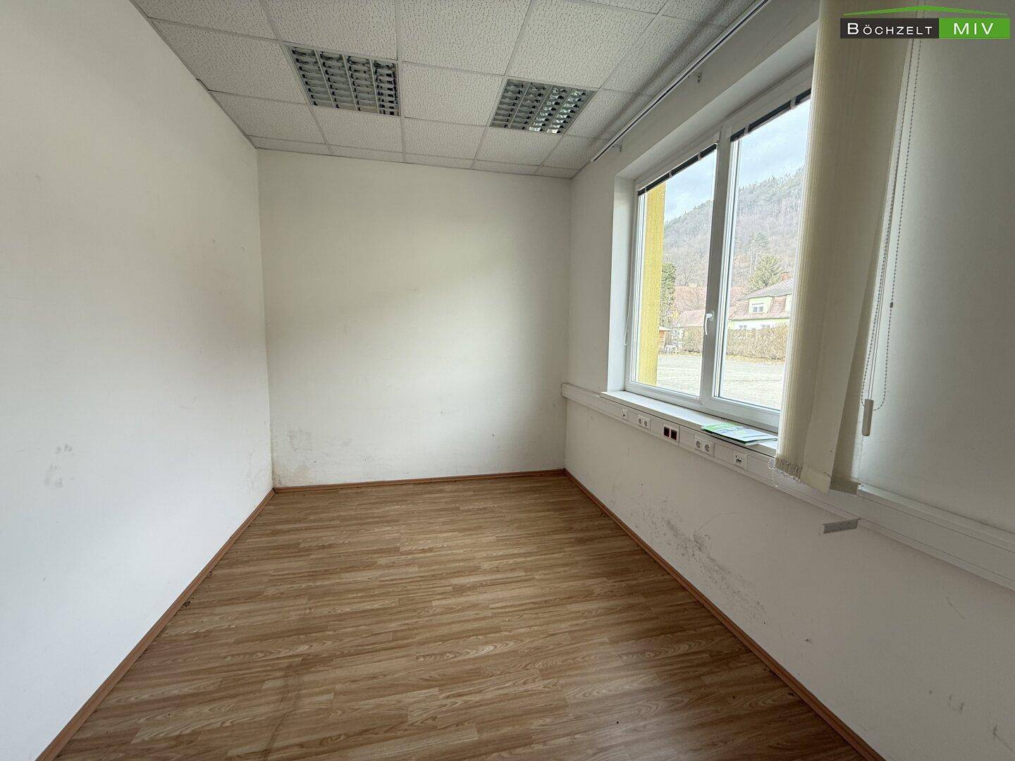 Leeres Büro mit Laminatboden und einem großen Fenster, das viel Tageslicht hereinlässt.