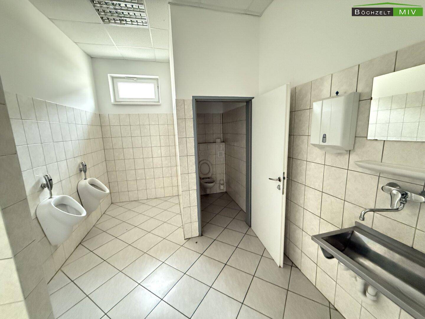 Sanitärbereich mit weißen Fliesen, zwei Urinalen, einem Waschbecken und einem kleinen Fenster.