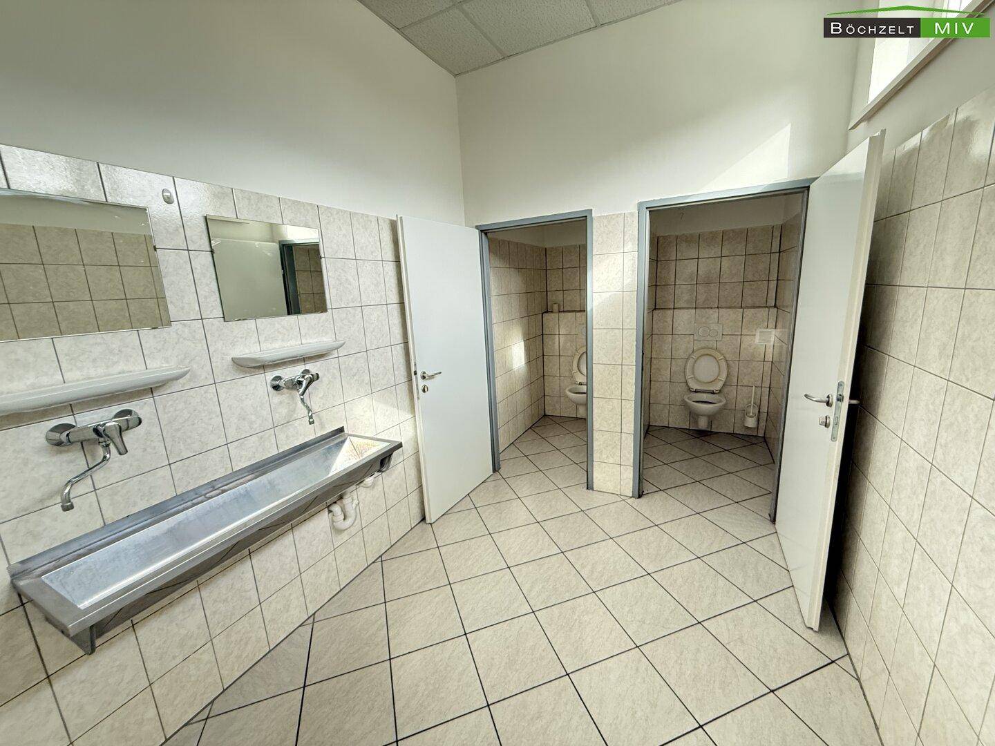 Sanitärraum mit weißen Fliesen, einem langen Waschbecken und mehreren Toilettenkabinen.