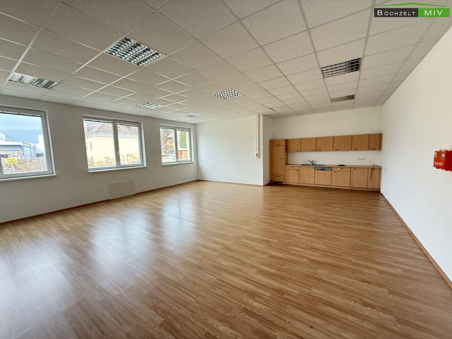 Geräumiger Raum mit Laminatboden, großen Fenstern und einer schlichten Küchenzeile an der Seite.