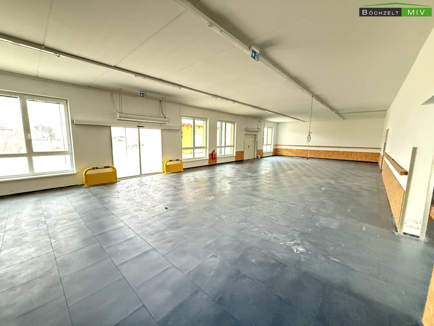 Weitläufiger, heller Raum mit dunklem Fliesenboden und großen Fenstern, die viel Tageslicht hereinlassen.