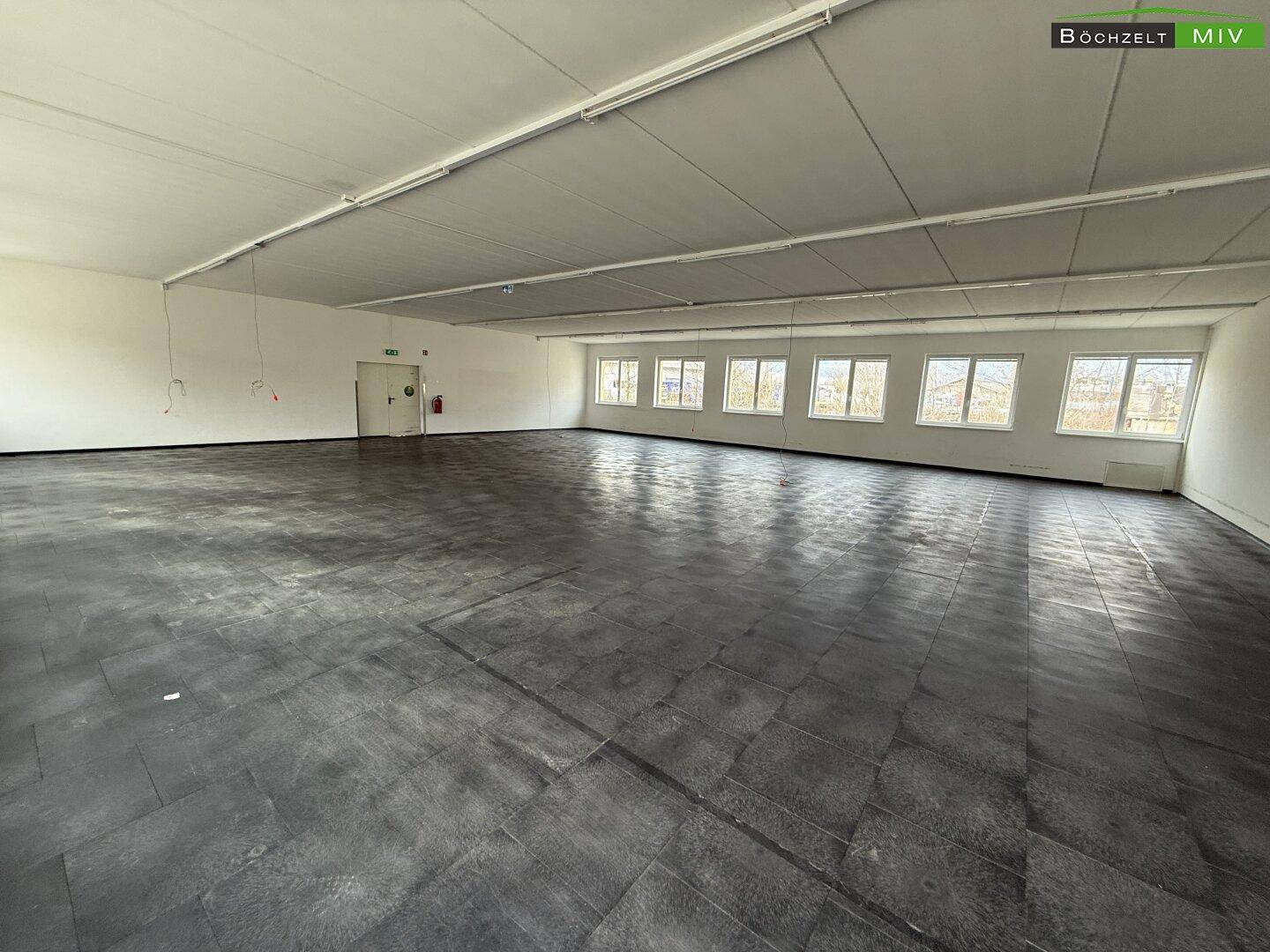 Weitläufiger, leerer Raum mit dunklem Fliesenboden und vielen Fenstern, ideal für flexible Nutzung.