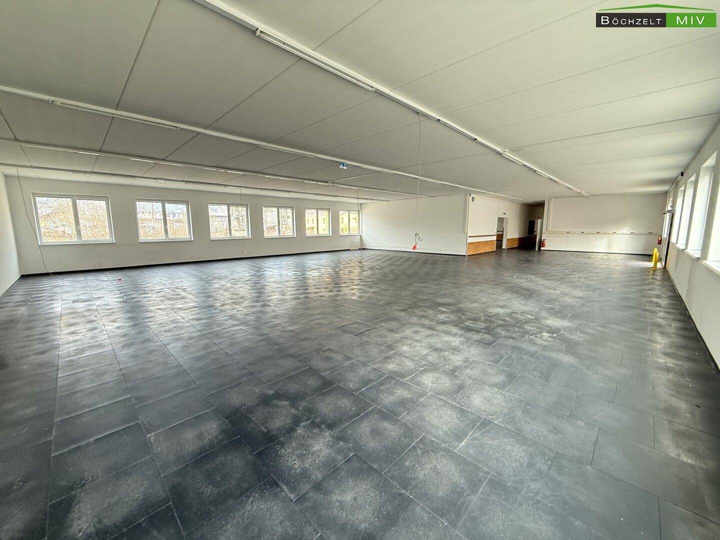 Geräumiger, heller Raum mit dunklem Fliesenboden und einer langen Fensterreihe, die den Raum belichtet.