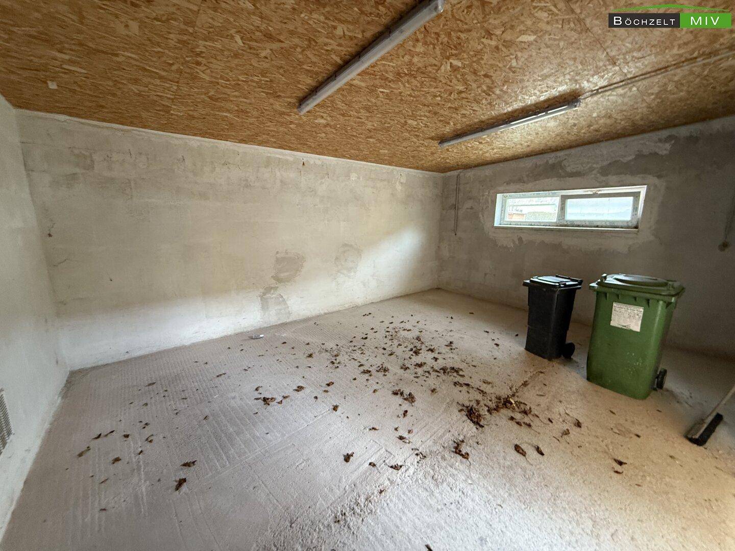 Leerer Abstellraum mit Betonboden, einem kleinen Fenster und sichtbaren Holzplatten an der Decke.