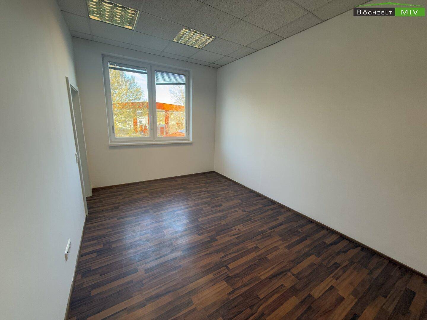 Heller Büroraum mit großem Fenster, das viel Tageslicht hereinlässt, und dunklem Laminatboden.