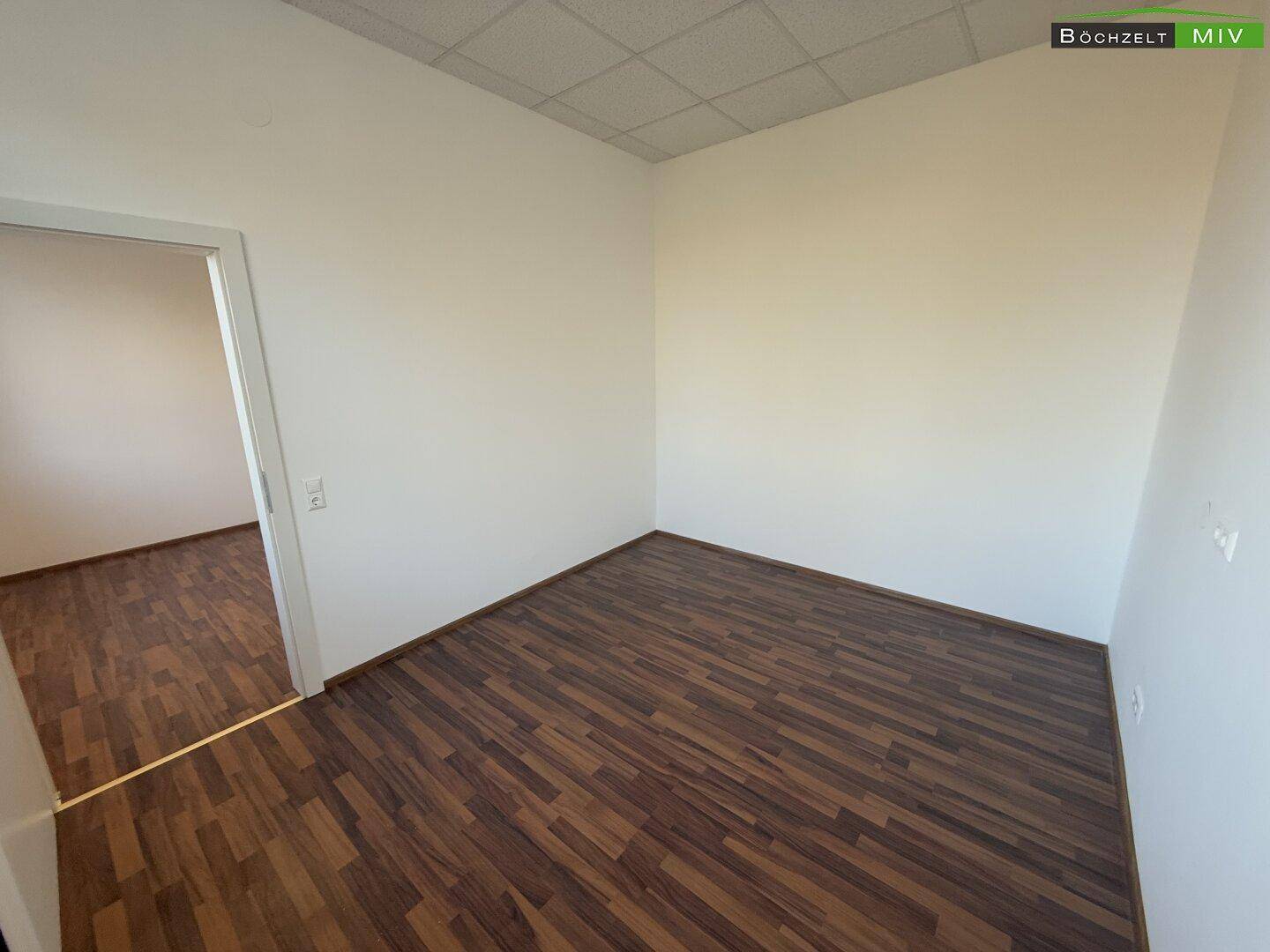 Heller Büroraum mit dunklem Laminatboden und Zugang zu einem weiteren Zimmer, ideal für flexible Nutzung.