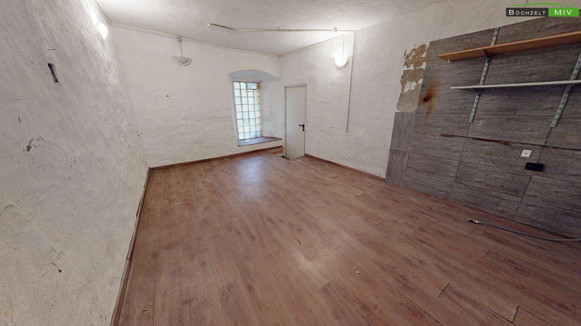 Geräumiger Raum mit Holzboden, weiß gestrichenen Wänden und einem Fenster mit Gitter.