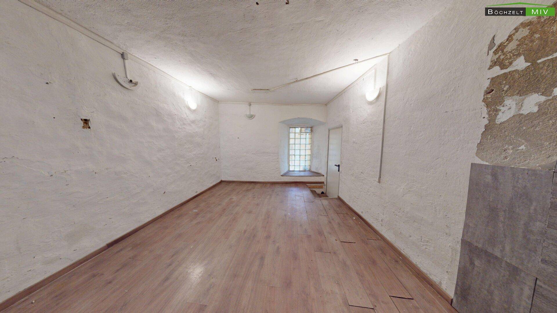 Heller Raum mit Holzboden, weiß getünchten Wänden und einem Fenster mit Gitter, gut beleuchtet.