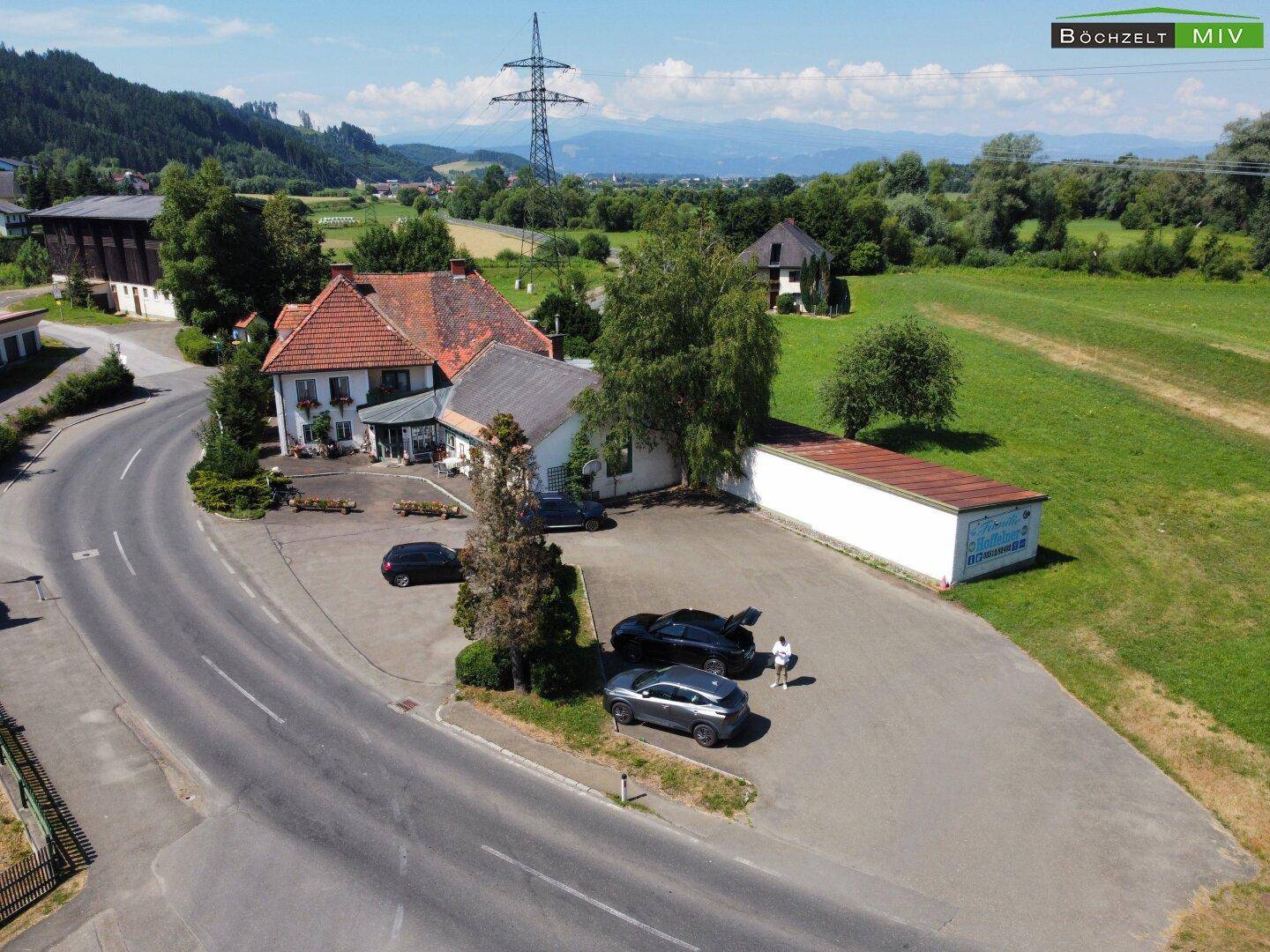 Luftaufnahme des Anwesens mit großem Parkplatz, einem Nebengebäude und der umliegenden grünen Landschaft.