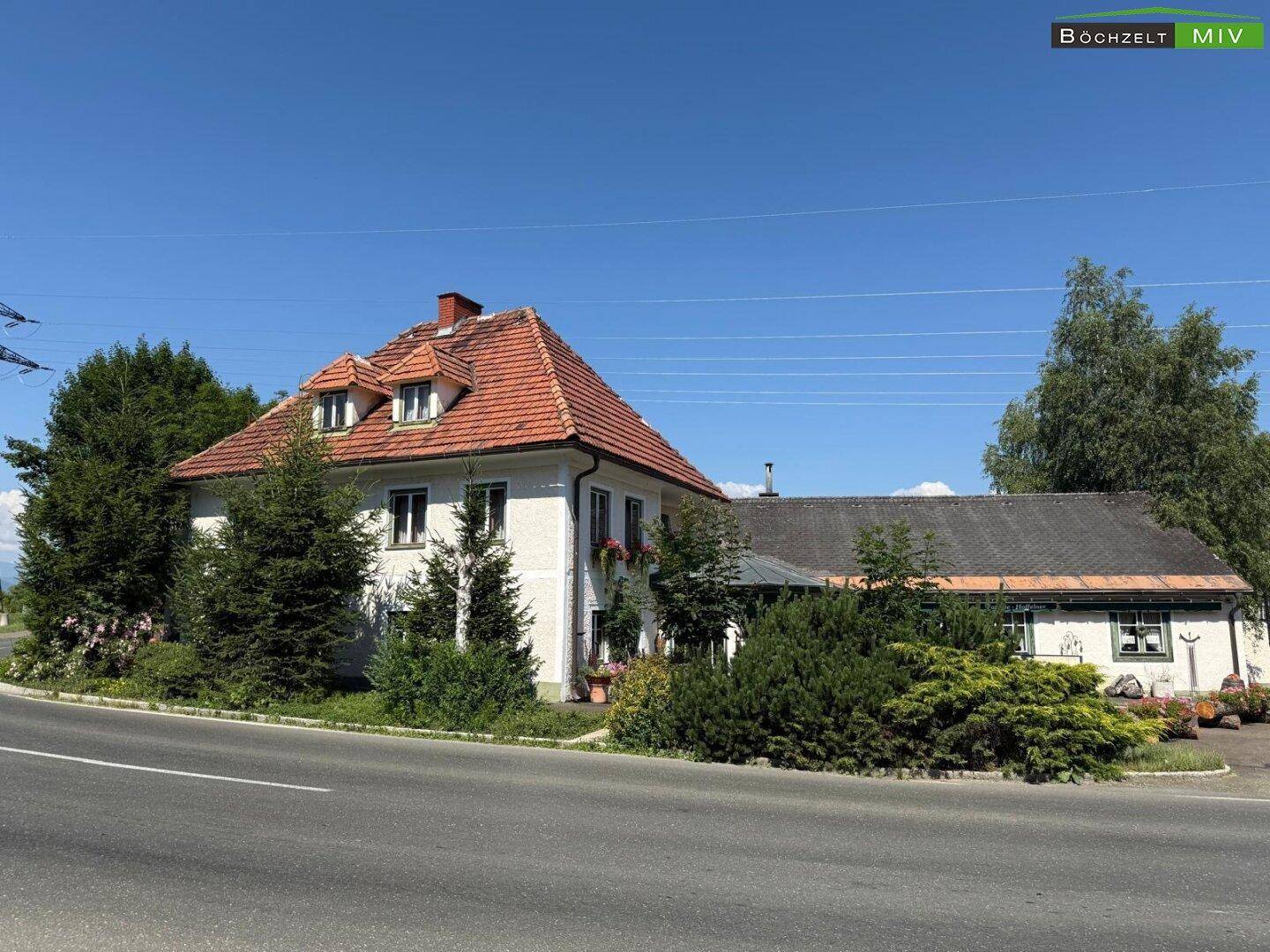 Vorderansicht des Anwesens von der Straße, umgeben von Bäumen und Sträuchern unter blauem Himmel.