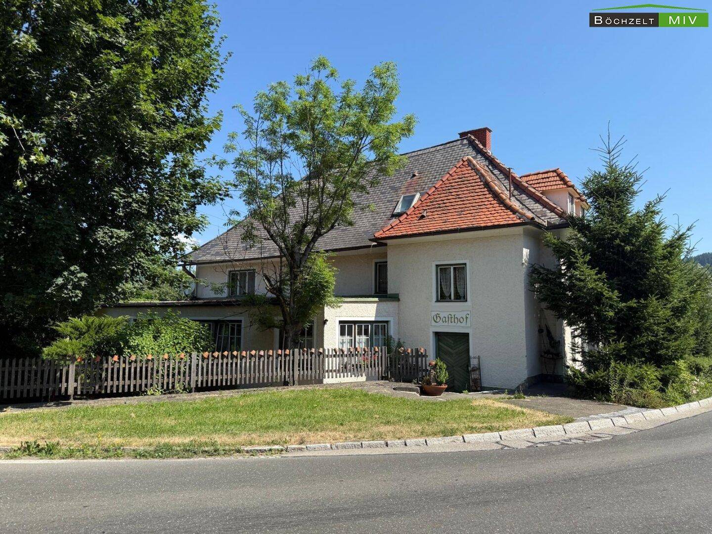 Seitenansicht des Gebäudes mit Gartenbereich, Holzzaun und einem Schild, das auf einen Gasthof hinweist.