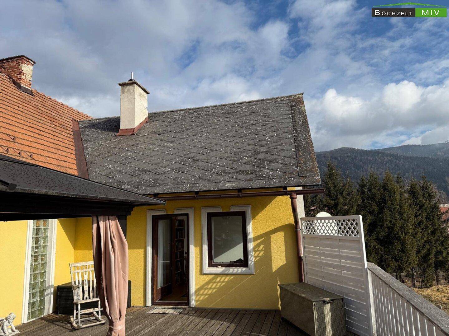 Teilansicht des Hauses mit Holzdielen-Terrasse und Blick auf die bewaldeten Berge.