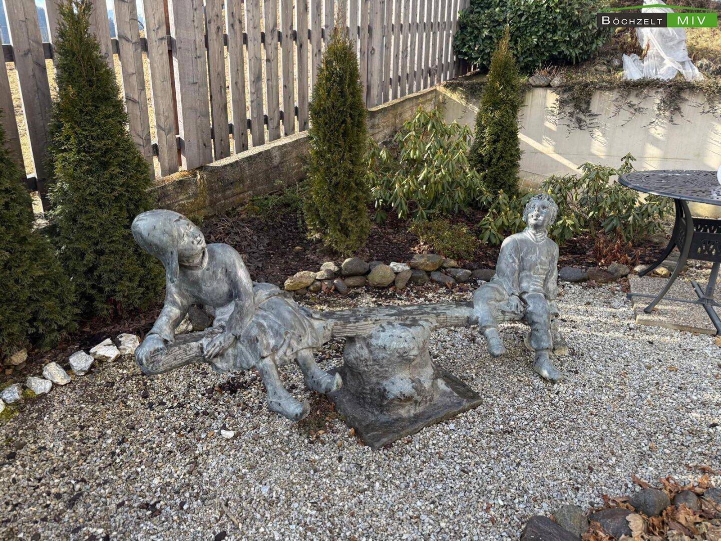 Gartendekoration mit einer Skulptur von Kindern auf einer Wippe in einem Kiesbeet.